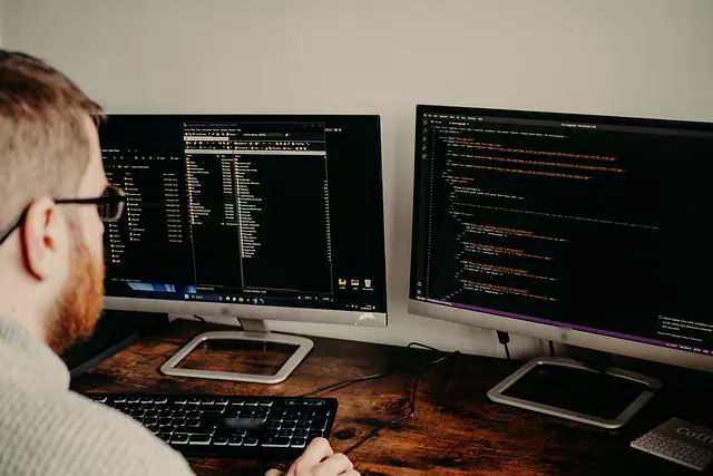 A developer seen from behind, working across two monitors of code.