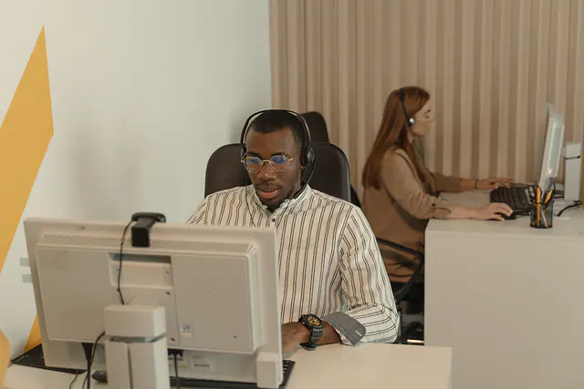 An engineer wearing a headset at a desk with a monitor, talking with a colleague.